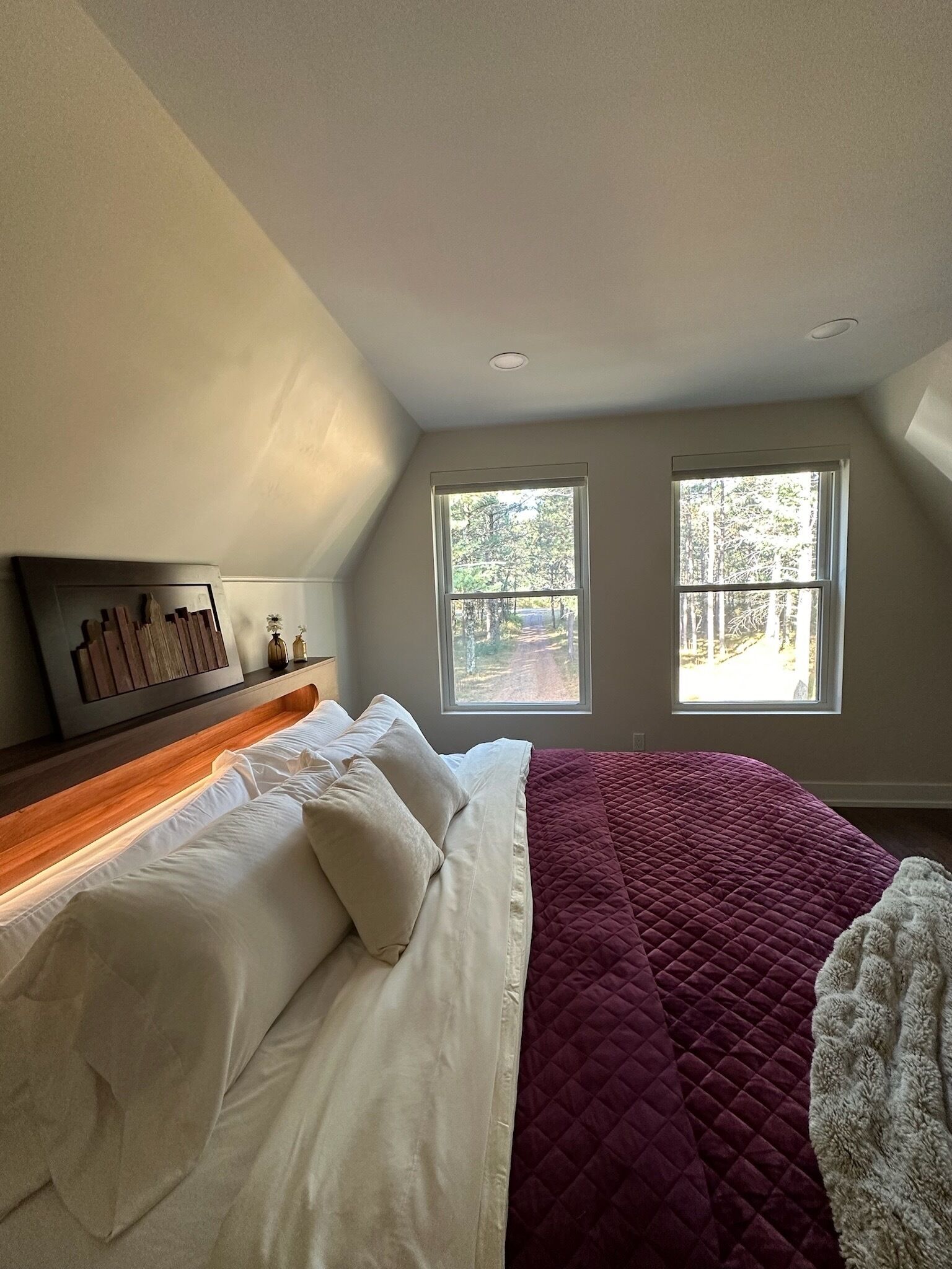 A bedroom with a bed, two windows, a headboard, and a view of trees.