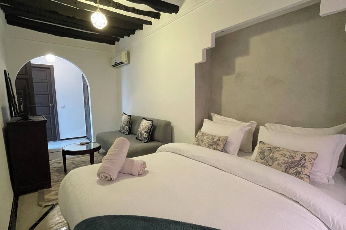 A bedroom with a large bed, a chair, a desk, and an arched doorway.