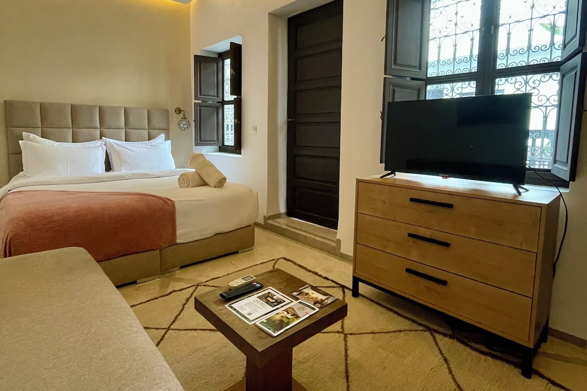 A hotel room with a bed, a sofa, a TV on a wooden cabinet, and a window with decorative iron bars.