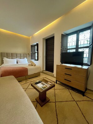 Habitación de hotel con cama, sofá, televisor sobre un mueble de madera y ventana con rejas decorativas de hierro.