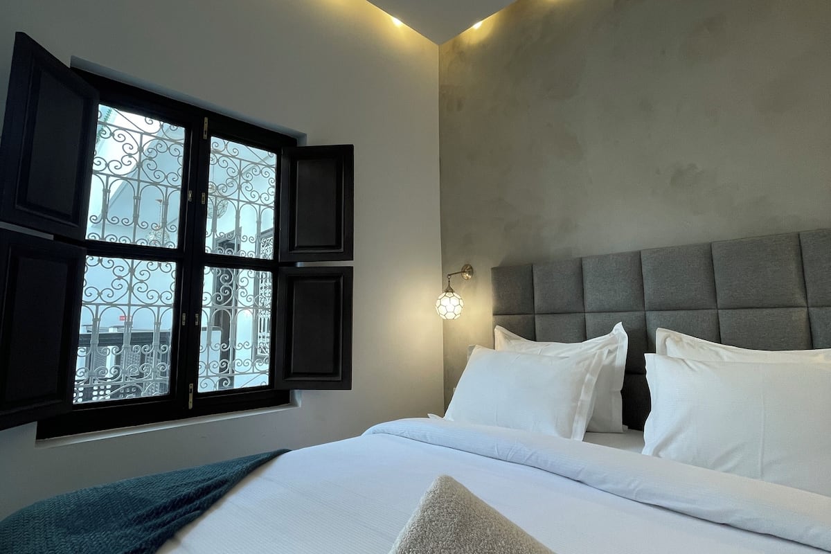 A bedroom with a bed, pillows, a window with a view, and a wall-mounted light fixture.