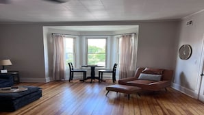 Una habitación con piso de madera, un sofá de cuero marrón, una mesa redonda con sillas y un ventanal con cortinas.
