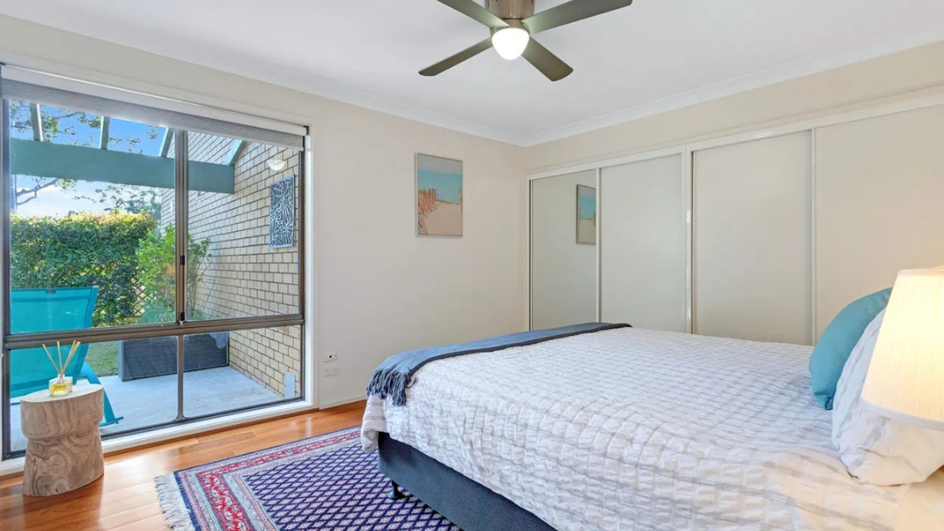 Un dormitorio con un amplio ventanal corredera a un balcón, una cama con cubrecama azul y blanco, un ventilador de techo y una pequeña mesita redonda con una vela.