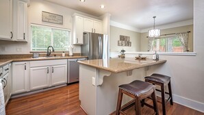 A kitchen with white cabinets, a stainless steel refrigerator, a granite countertop, and two stools. - Serenity Awaits: Pine Trees Retreat w/ AC and EV (Flagstaff)