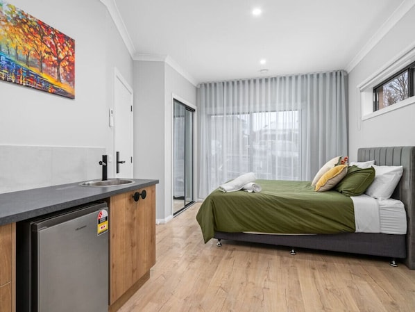 A modern bedroom with a bed, a small kitchenette, and a sliding glass door to a balcony.