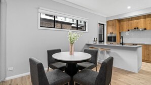 A modern kitchen with a central island, a dining area with a round table, and a window with a view of a brick building. - New Central 2 bedroom townhouse! (Beechworth)