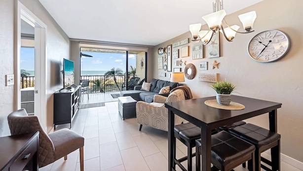 A modern living room with a sofa, dining table, and chairs. There is a TV, a chandelier, and a clock on the wall. - Beachfront paradise in this gorgeous renovated ground floor condo! (Marco Island)