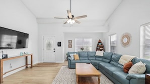 A living room with a sectional sofa, a wooden coffee table, a ceiling fan, and a TV on the wall. - Beach Getaway w Pool | Good Morning Rentals (Jacksonville Beach)