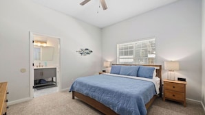 A bedroom with a bed, bedside tables, a ceiling fan, a window with blinds, and a door leading to a bathroom. - Beach Getaway w Pool | Good Morning Rentals (Jacksonville Beach)