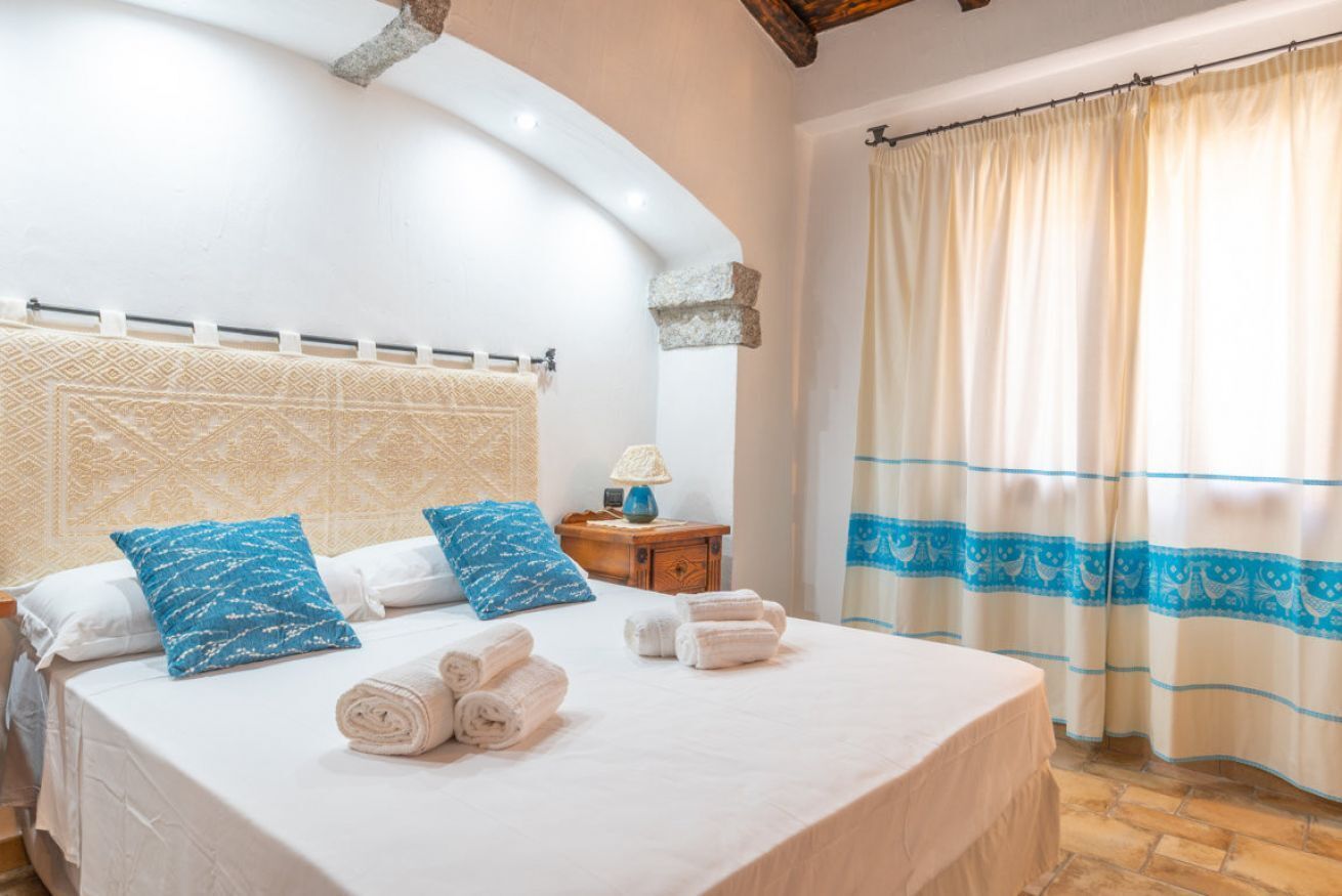 A neatly made bed with white linens and blue patterned pillows, a small wooden nightstand with a lamp, and a window with a beige curtain.