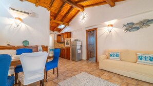 A cozy living room with a wooden ceiling, a beige sofa with patterned cushions, a dining table with blue chairs, and a kitchen in the background. - PV9: One-Bedroom Apartment with Romantic Terrace in the City Center (Olbia)