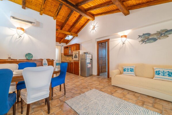 Una sala de estar acogedora con techo de madera, un sofá beige con cojines estampados, una mesa de comedor con sillas azules y una cocina al fondo.