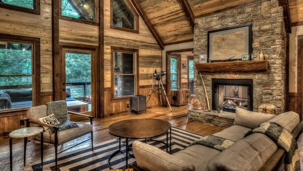 Interior rústico de una cabaña con chimenea de piedra, vigas de madera y ventanas amplias.