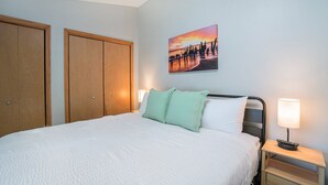 Una cama bien tendida con un edredón blanco, dos almohadas, una mesita de noche con una lámpara y un cuadro de un atardecer en la pared.