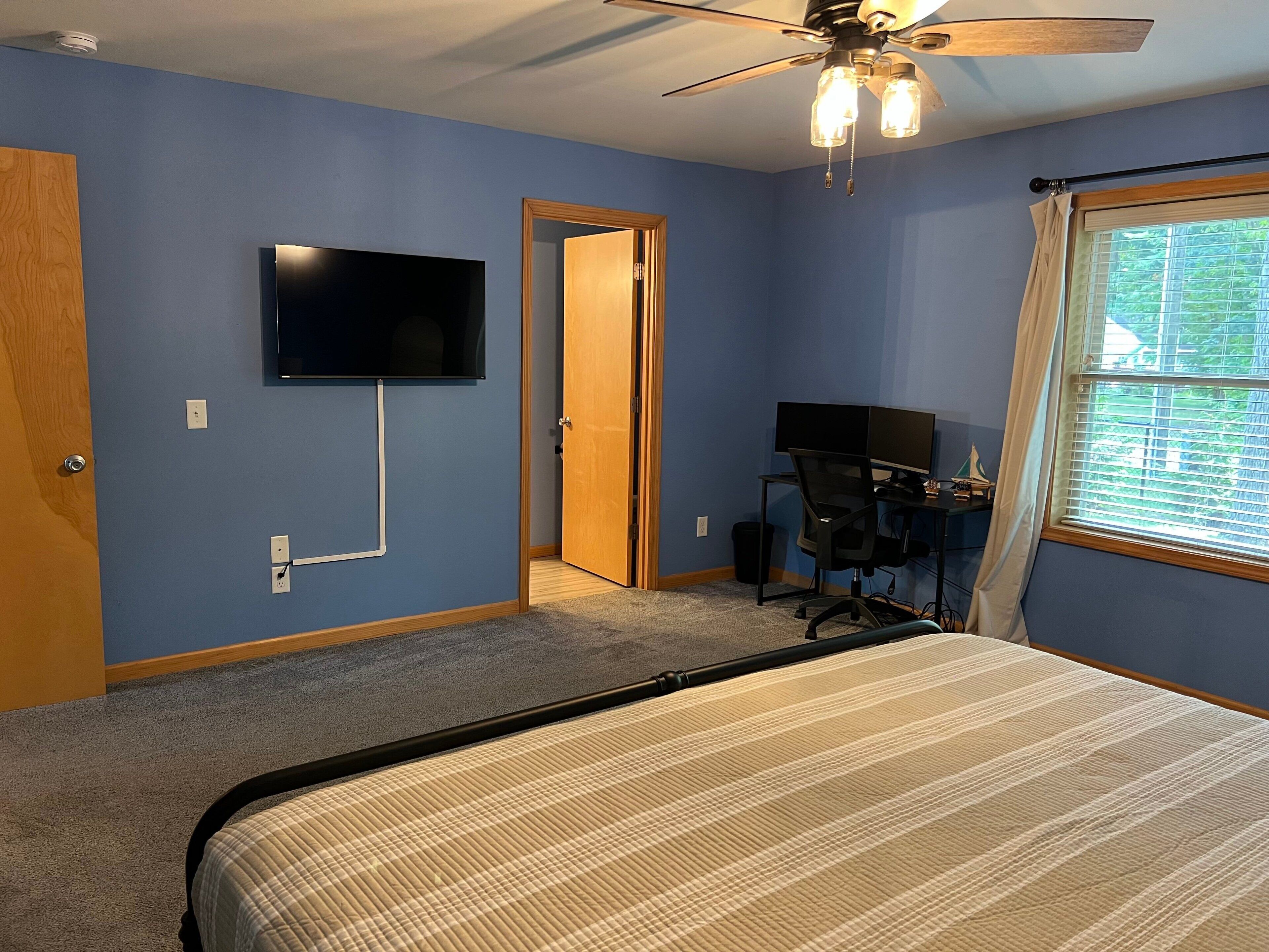 A bedroom with a bed, a desk with a computer, a TV on the wall, and a ceiling fan.
