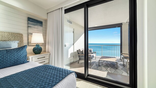 A bedroom with a large bed, a nightstand, a lamp, and a view of a balcony with outdoor furniture. - Summer House 904B | Oceanfront | Pools, Hot Tub, Tennis | My Beach Getaways (Orange Beach)