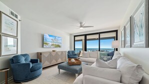 A modern living room with a view of the ocean, a ceiling fan, and a large window. - Summer House 904B | Oceanfront | Pools, Hot Tub, Tennis | My Beach Getaways (Orange Beach)