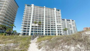 A multi-story hotel with balconies, a sandy pathway leading to the entrance, and palm trees in the background. - Summer House 904B | Oceanfront | Pools, Hot Tub, Tennis | My Beach Getaways (Orange Beach)
