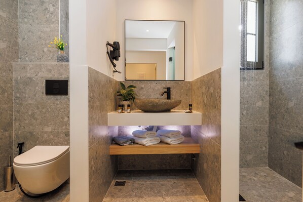 A modern bathroom with a stone sink, a mirror, a toilet, and a shelf with towels. - Soho SeaFront Suites (Hersonissos)