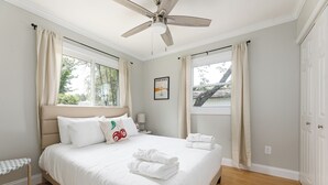 A bedroom with a bed, a ceiling fan, two windows with curtains, a nightstand, and a wall-mounted picture.