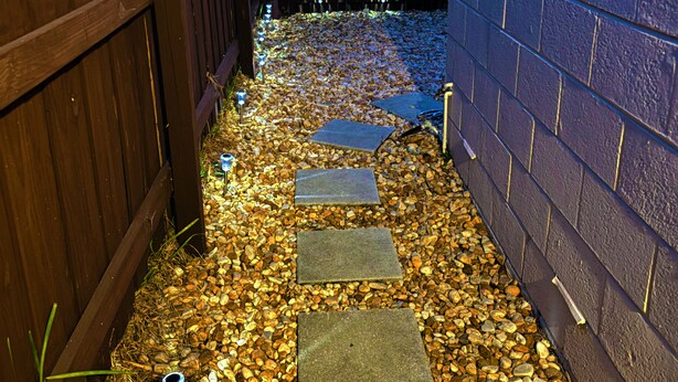 Un sentier pavé de pierres plates mène à une porte en bois, qui est elle-même entourée d’un mur de briques et de palmiers.