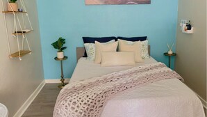 Une chambre à coucher avec un lit, une étagère fixée au mur ornée de plantes, une petite table avec une plante et un tableau représentant un coucher de soleil sur l’océan.