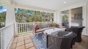 Un balcon avec un canapé, des fauteuils en osier, une table ornée d’un vase, et une vue sur les arbres et les bâtiments.