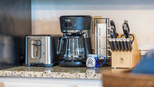 Una encimera de cocina con cafetera, tostadora, taza y bloque de cuchillos.
