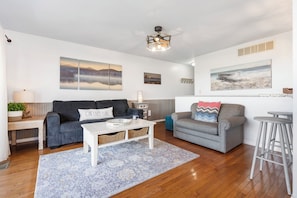 A living room with a sofa, armchair, coffee table, and a bar stool. There are wall art pieces and a ceiling light fixture.