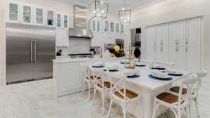 A modern kitchen with white cabinetry, stainless steel appliances, and a dining area with white chairs and a marble countertop. - Reunion's Finest | Luxury 10BR Home w Golf Course View, Private Pool, Theater and Arcade Near Disney (Kissimmee)