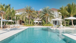 A pool area with lounge chairs and umbrellas, surrounded by palm trees and a building with a tiled roof.