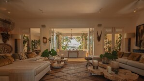 A spacious living room with a large sofa set, a coffee table, and a patterned rug. There are several potted plants and a large window providing a view of the outdoors.