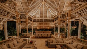 A spacious, open-air lounge with high ceilings and a thatched roof, featuring comfortable seating areas and a central bar.
