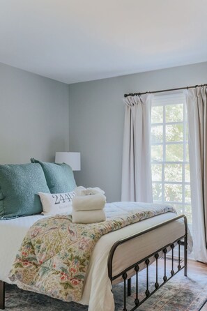 A bedroom with a bed, a window with curtains, a bedside lamp, and a floral bedspread. - Stuart Cottage: Walk to downtown + Sleeps 8 (Staunton)