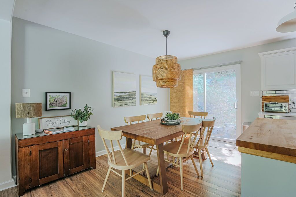 Un coin repas avec une table et des chaises en bois, un luminaire suspendu et une vue sur l’extérieur par une fenêtre.