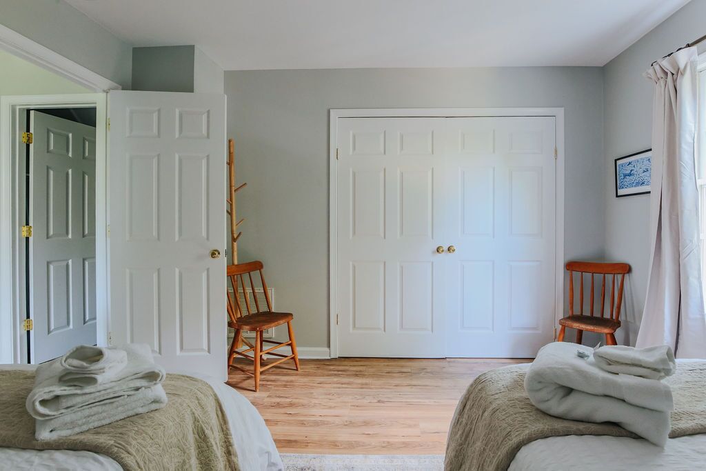 Une pièce avec deux lits, une chaise en bois, une porte blanche et un tableau encadré au mur.