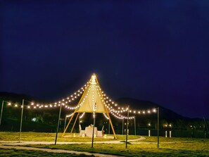 Uma tenda com luzes de corda, uma mesa e cadeiras montadas em um campo aberto à noite.