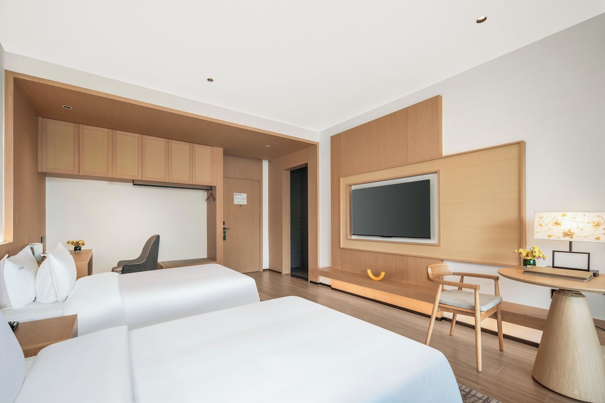 Quarto de hotel moderno com duas camas, uma televisão, uma mesa de escritório com cadeira e uma mesinha de cabeceira com abajur.