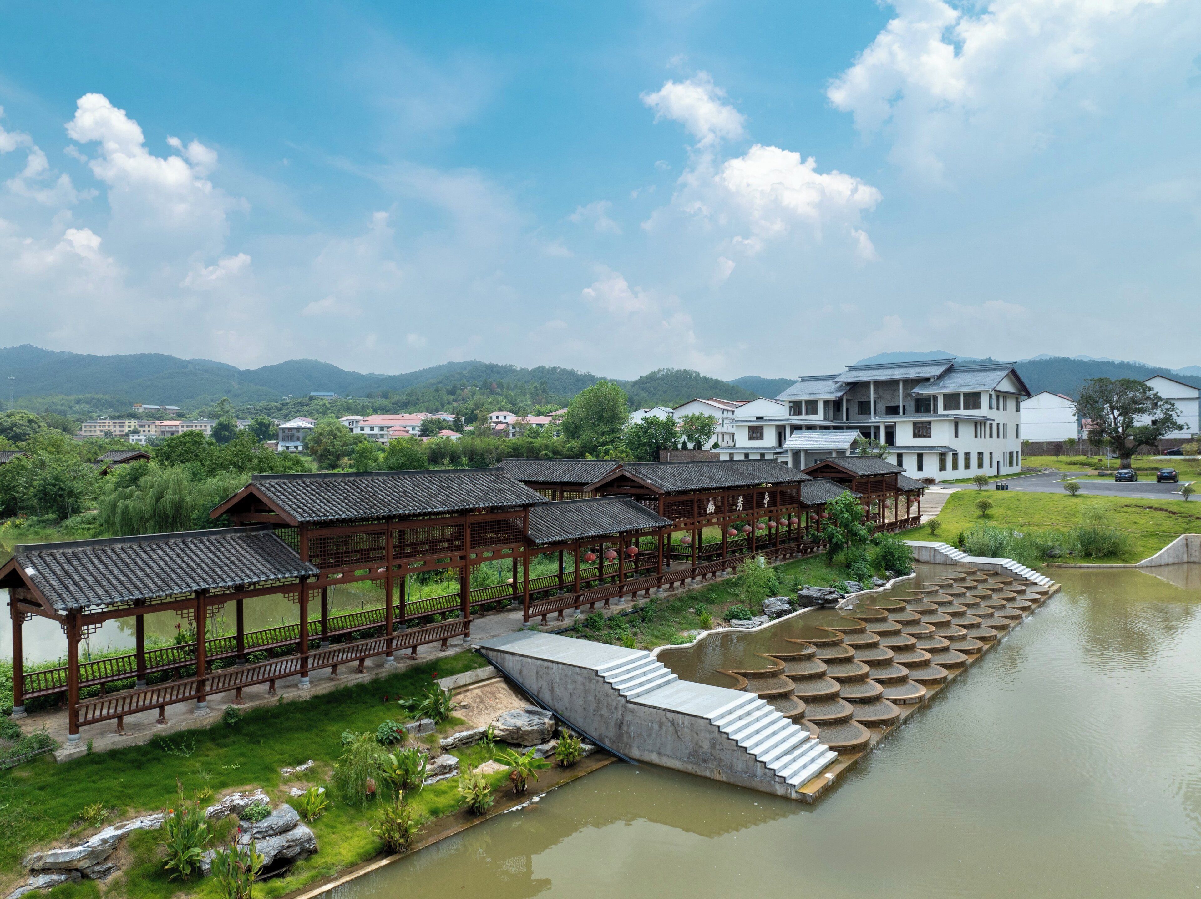 A traditional wooden pavilion with a stone bridge over a pond, surrounded by lush greenery and modern buildings in the background.