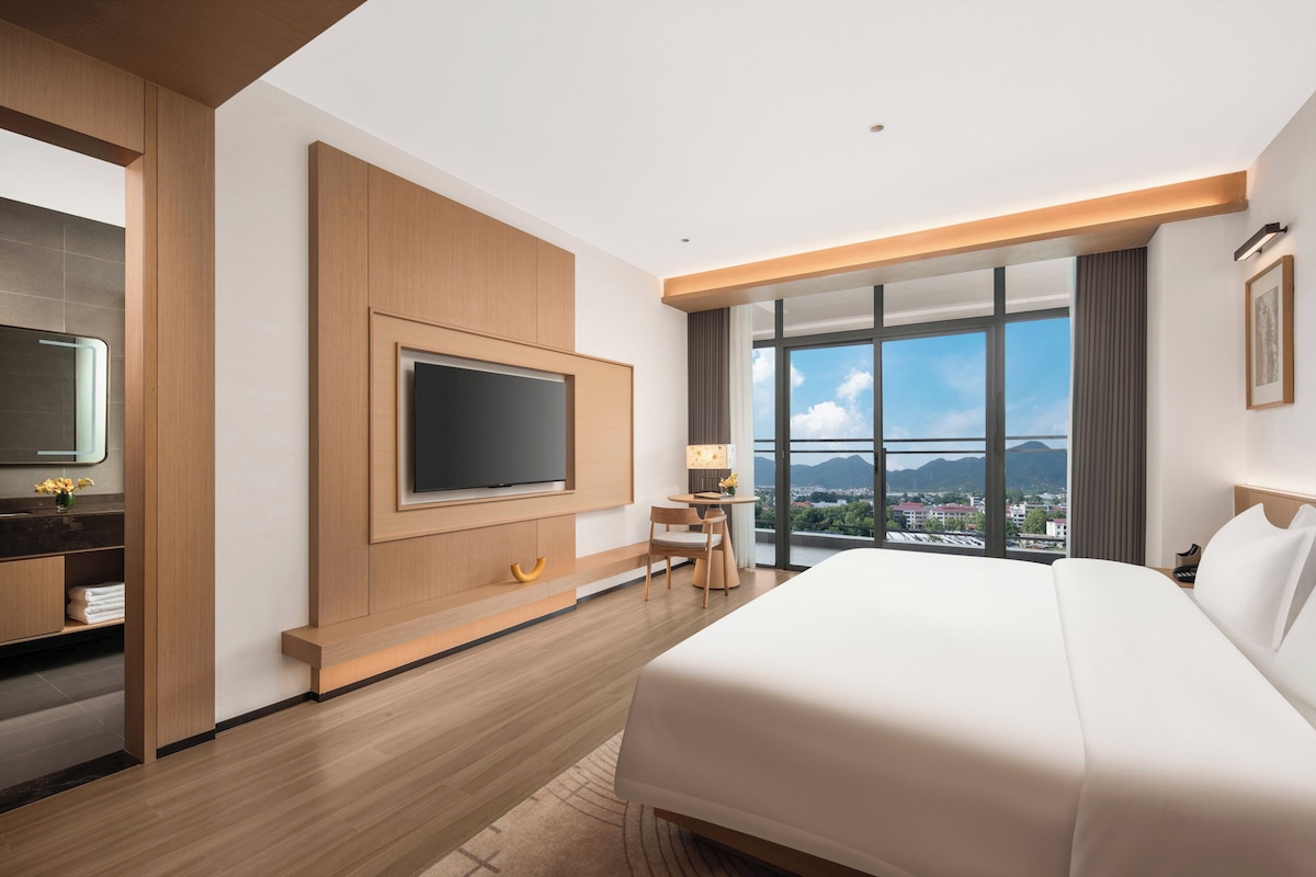 Quarto de hotel moderno com uma cama grande, televisão de tela plana na parede, uma mesa pequena com uma cadeira e vista da cidade através de grandes janelas.