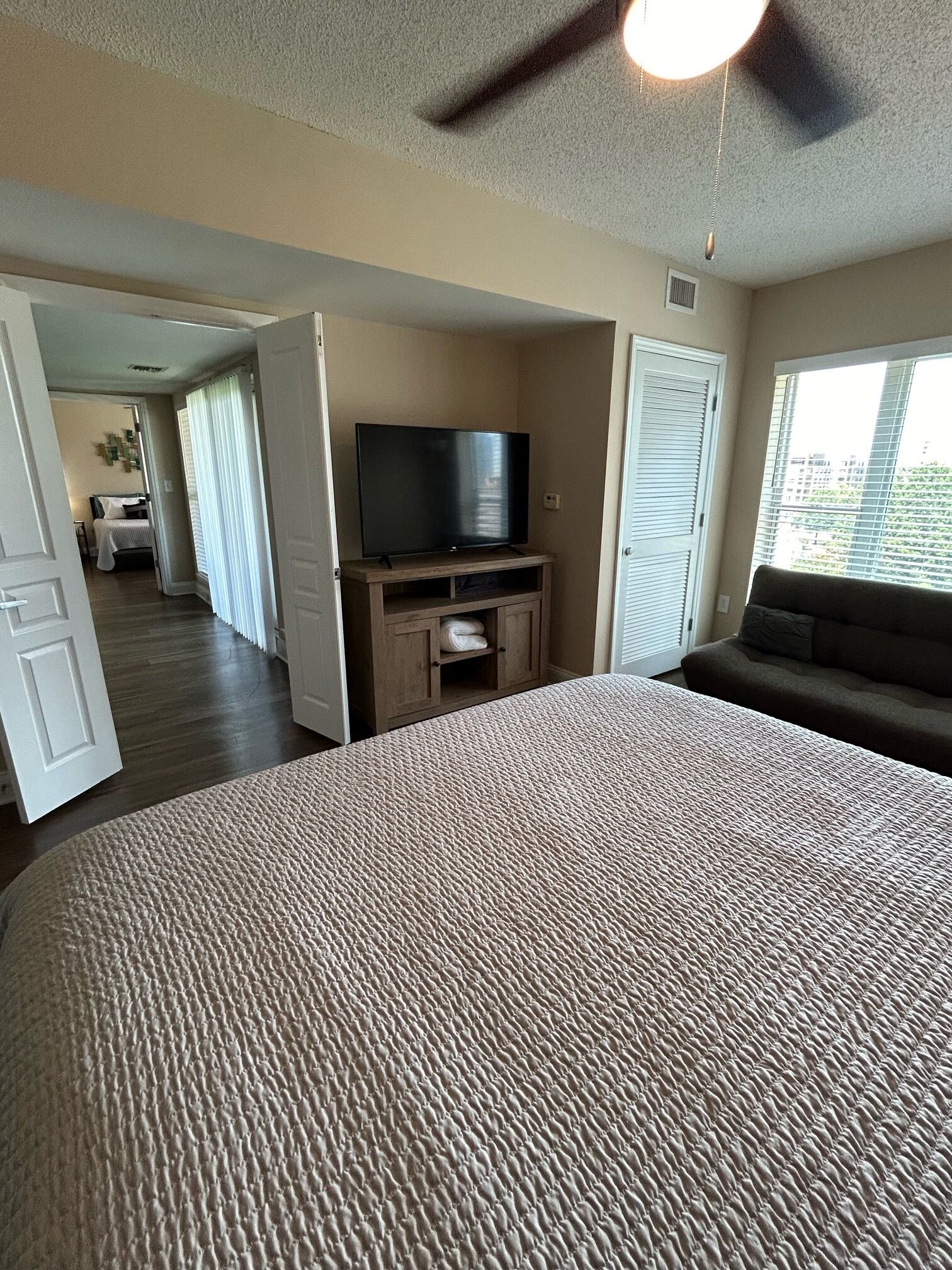 A hotel room with a large bed, a television on a wooden stand, a sofa, and a view of the city through the window.