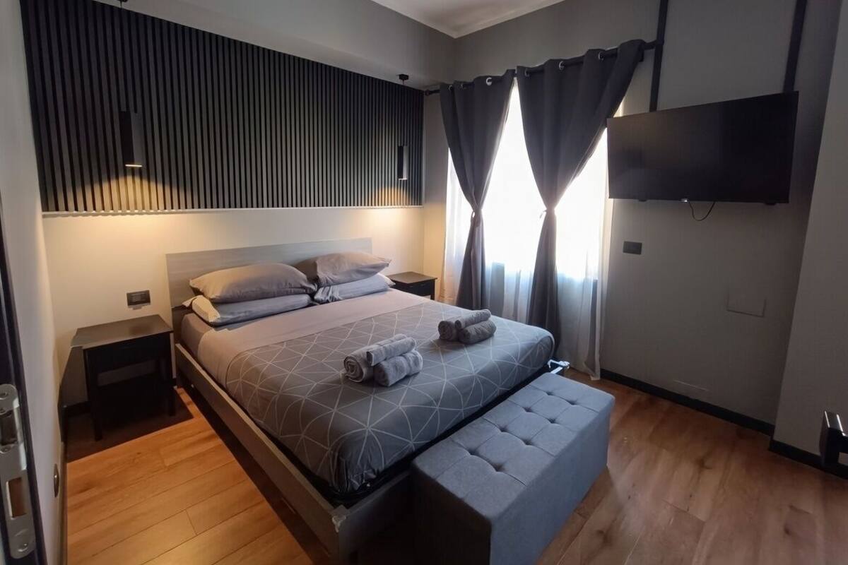 A modern bedroom with a large bed, a bedside table, a bench, a TV mounted on the wall, and a window with curtains.