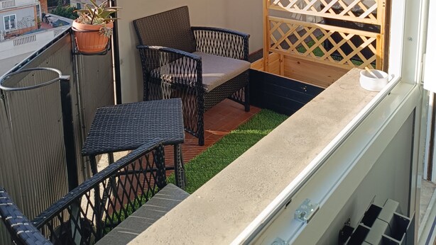 Ein Balkon mit Korbmöbeln, einer Topfpflanze und Blick auf eine Stadtlandschaft.