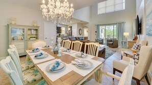 A dining area with a wooden table set for six, surrounded by white chairs with floral upholstery. A large chandelier hangs from the ceiling, and a kitchen is visible in the background.