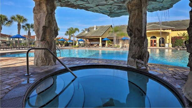 A pool area with a hot tub, palm trees, and a building with a tiled roof in the background.