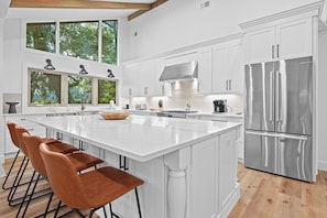 A modern kitchen with white cabinetry, a large island with bar stools, stainless steel appliances, and large windows offering a view of greenery.