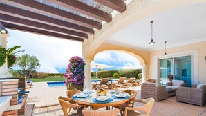 An outdoor patio with a round table set for dining, surrounded by wicker chairs, and overlooking a swimming pool. - Villa Monte Rei Golf (Vila Real Santo Antonio)