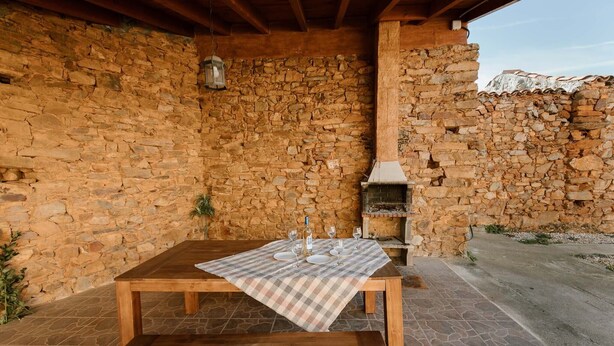 Un área de comedor al aire libre con paredes de piedra, una mesa de madera preparada para dos comensales, un banco y una chimenea de piedra.