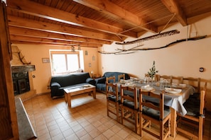 Ein gemütlicher Wohnbereich mit Kamin, einer Couch, einem Esstisch für vier Personen und einer Holzbalkendecke.
