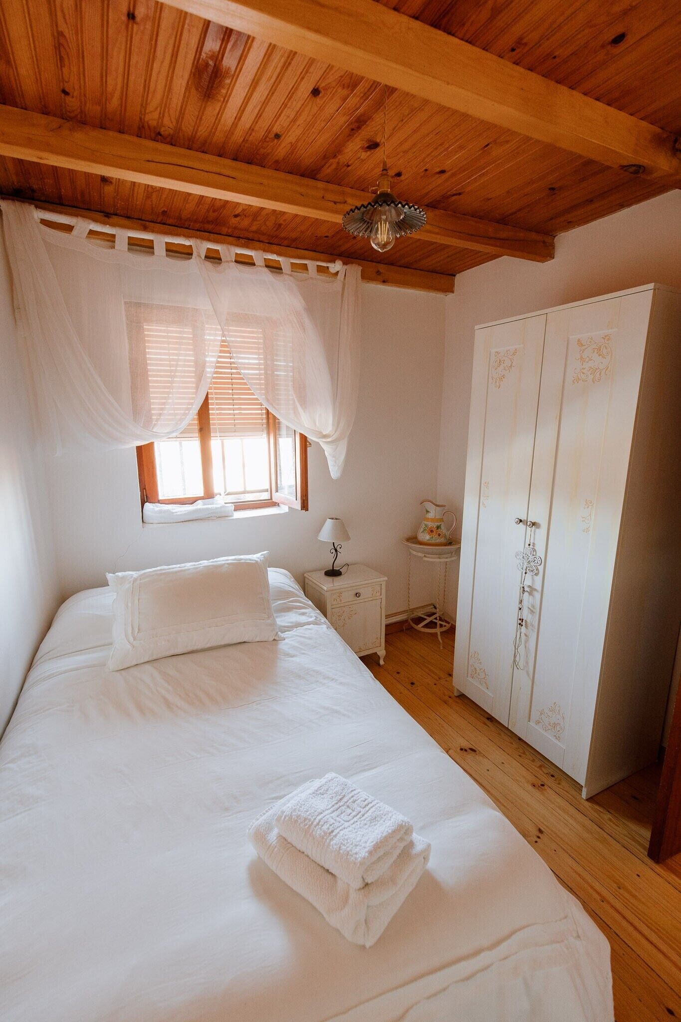 A bedroom with a wooden ceiling, a bed with white linens, a window with sheer curtains, a bedside table with a lamp, and a wardrobe.